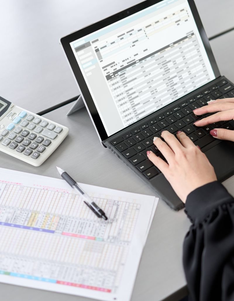 Person entering data on a laptop beside printed spreadsheets